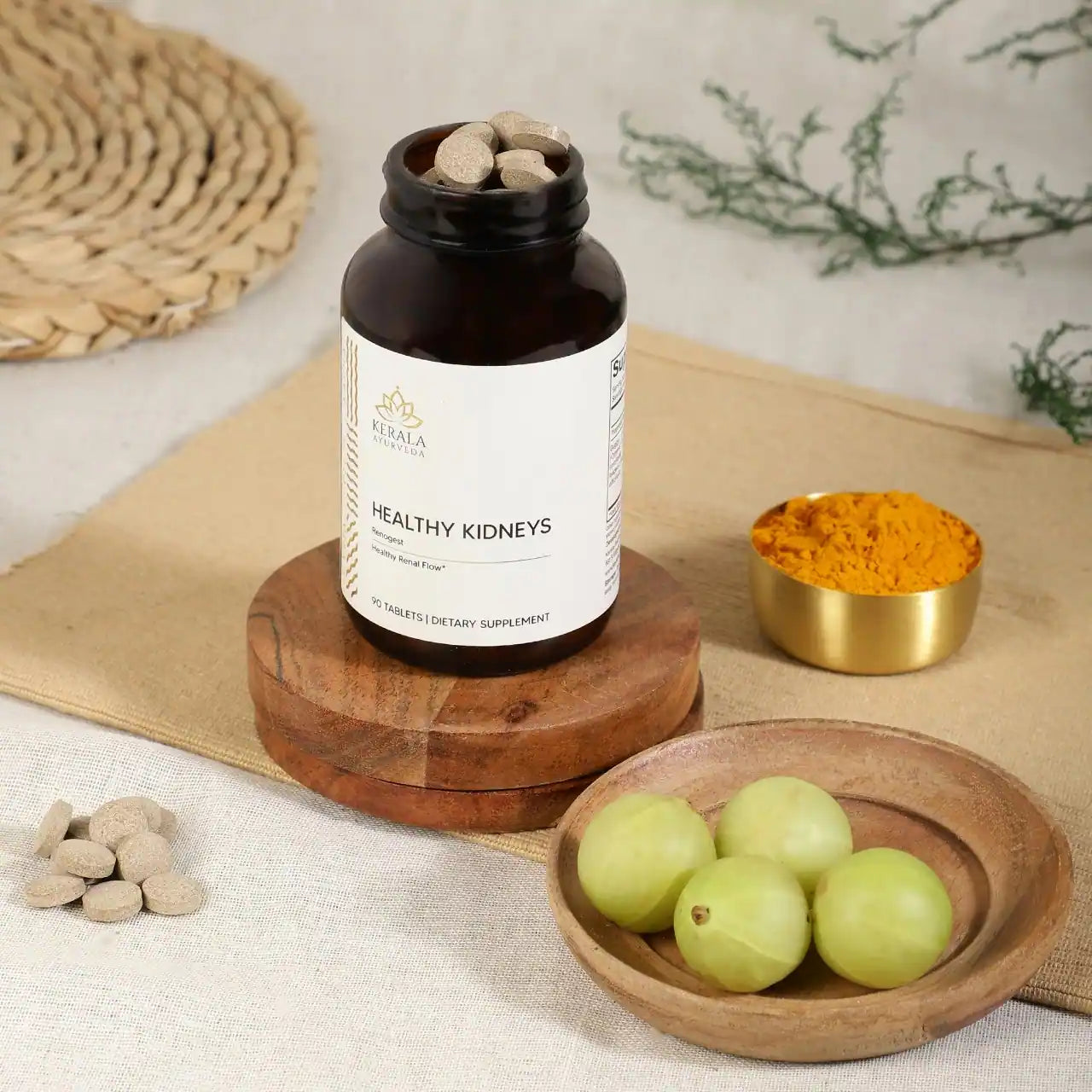 A open bottle of Healthy Kidneys dietary supplement with 90 tablets, labeled as Renogest, by Kerala Ayurveda. The bottle is places in a beige background with turmeric and gooseberries lying around