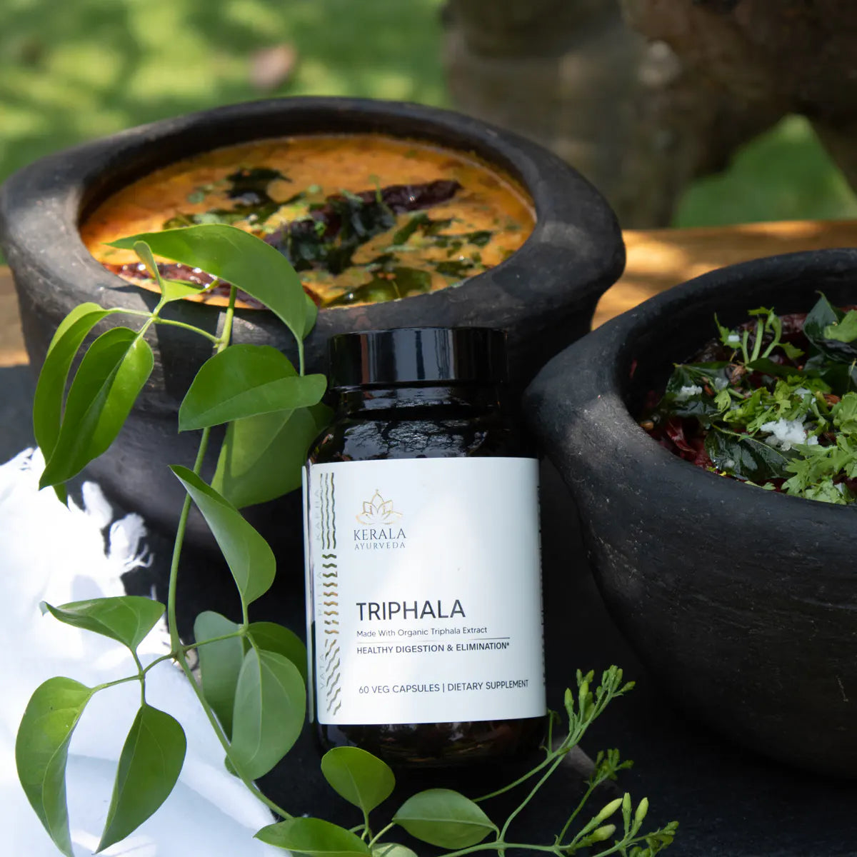 A bottle of Triphala dietary supplement next to two bowls of food, surrounded by fresh herbs and plants.
