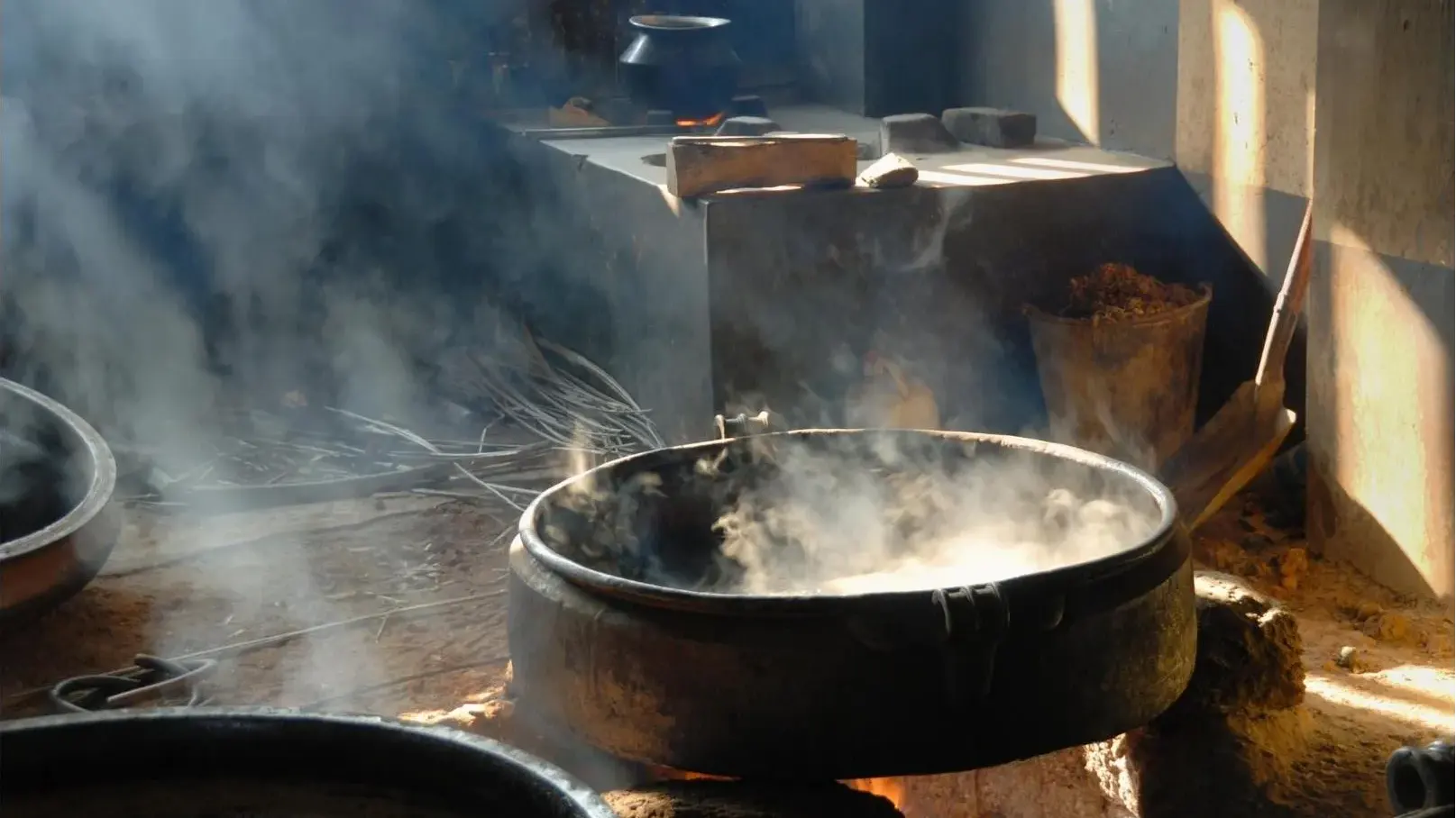 A traditional cooking pot with steam rising, in a rustic kitchen filled with smoke and natural light.