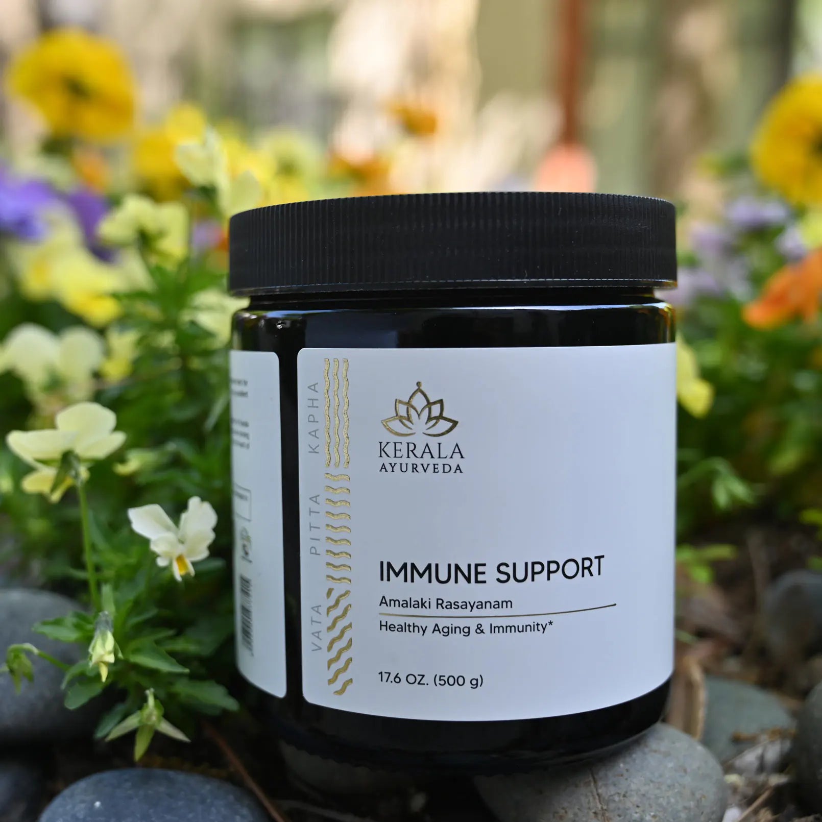 Black jar of Kerala Ayurveda Immune Support supplement surrounded by colorful flowers and stones.