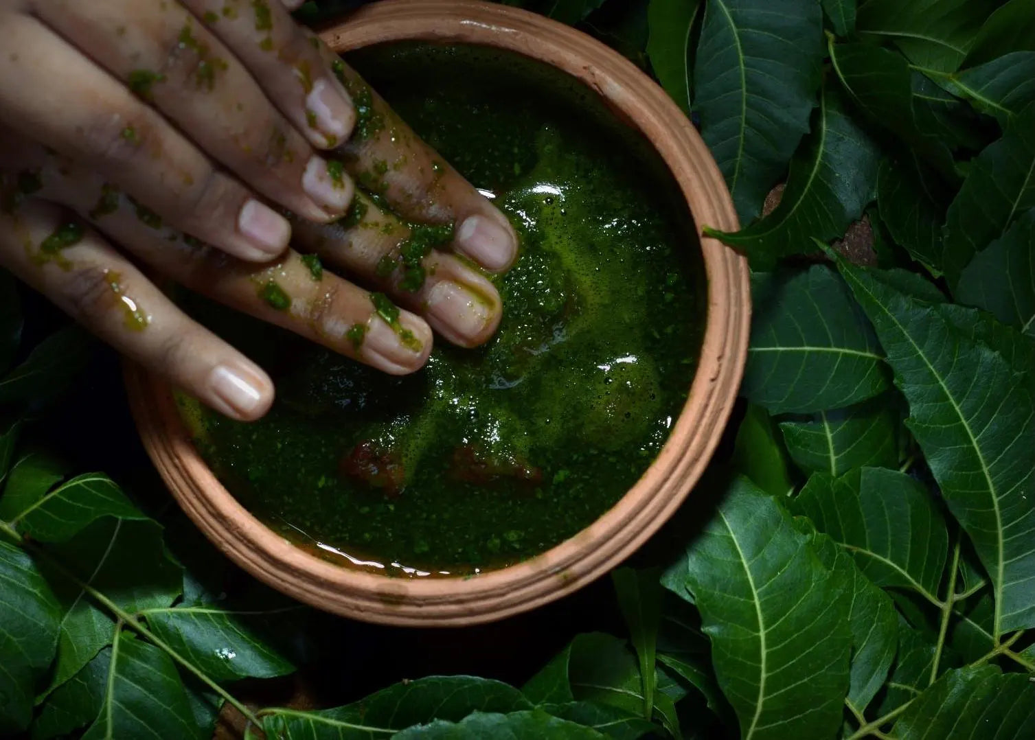 A hand with green paste in a clay bowl surrounded by fresh green leaves.