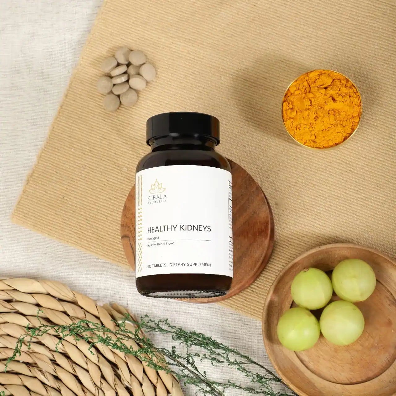 A bottle of Healthy Kidneys dietary supplement with 90 tablets, labeled as Renogest, by Kerala Ayurveda. The bottle is places in a beige background with turmeric, capsules and gooseberries lying around